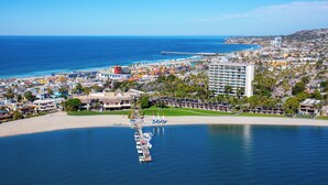 Vista aérea de una ciudad costera con un puerto deportivo, hoteles frente a la playa y un hotel destacado con un amplio balcón con vista al océano.