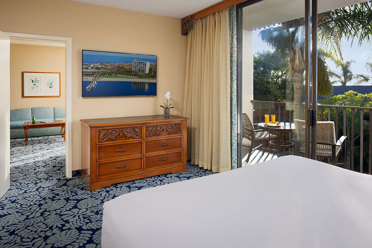 Quarto de hotel com uma cama grande, cómoda de madeira, quadro pendurado na parede, varanda com mesa e cadeiras, e vista para o exterior.