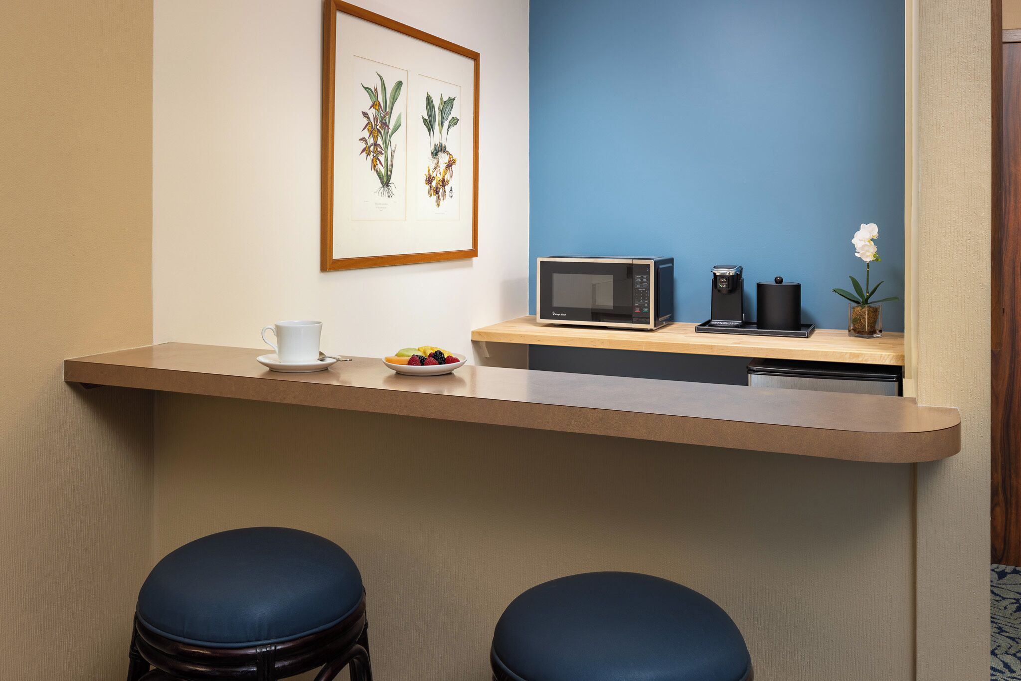 Un espace d’accueil d’hôtel avec un comptoir en bois, deux tabourets bleus rembourrés, une estampe botanique encadrée, un four à micro-ondes, une machine à café et une petite orchidée en pot.