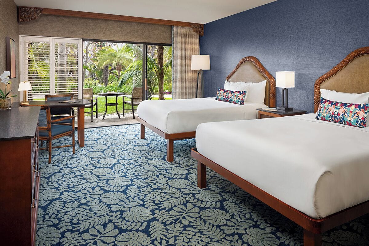 A hotel room with two beds, a desk, and a view of a garden.