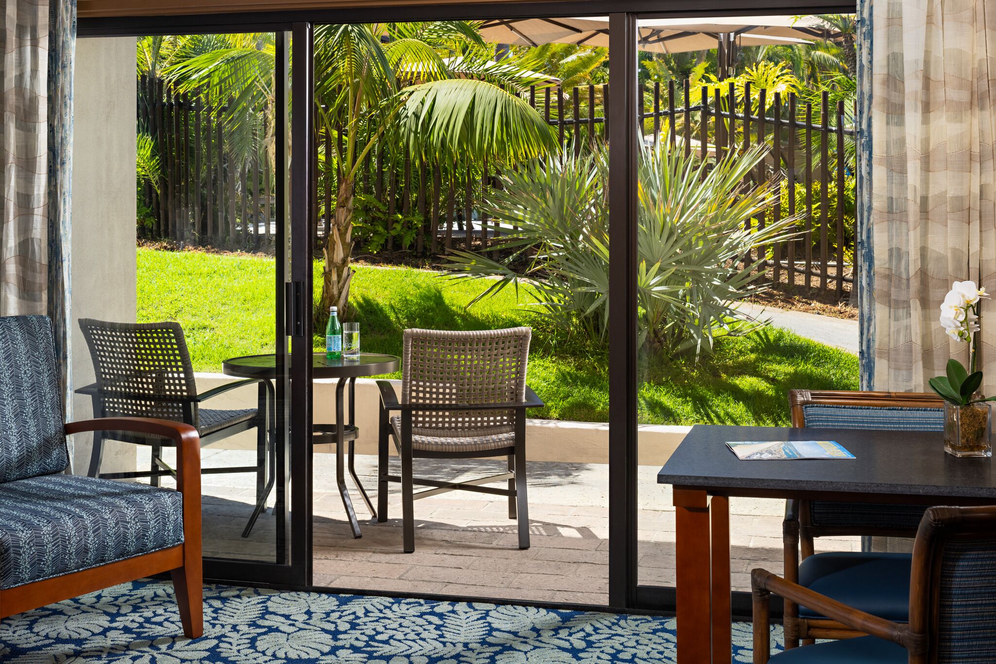 Une pièce dotée d’une porte-fenêtre coulissante donnant sur une terrasse aménagée avec des chaises, une table et une vue sur un jardin luxuriant.