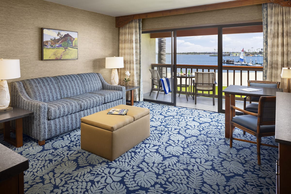 A hotel room with a sofa, ottoman, and a table with chairs. There is a balcony with a view of the water and boats.