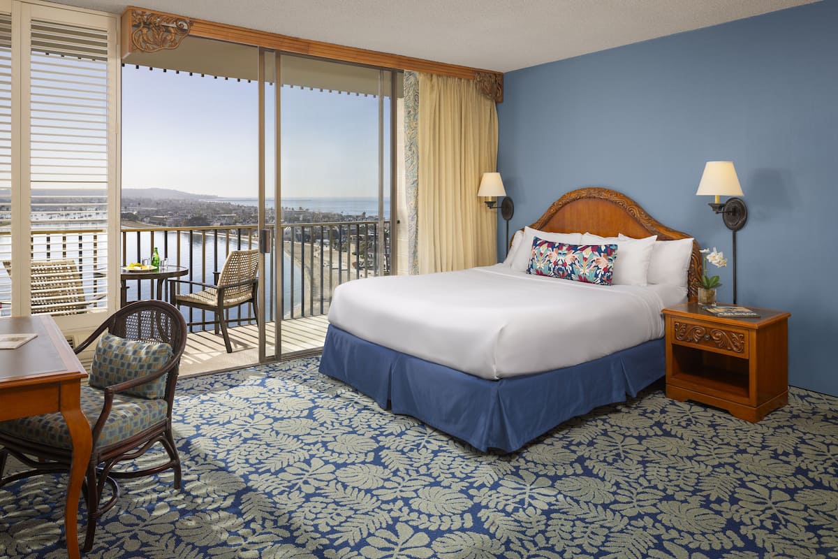 A hotel room with a large bed, two bedside lamps, a chair, a small table, and a view of the city and water through a sliding door.