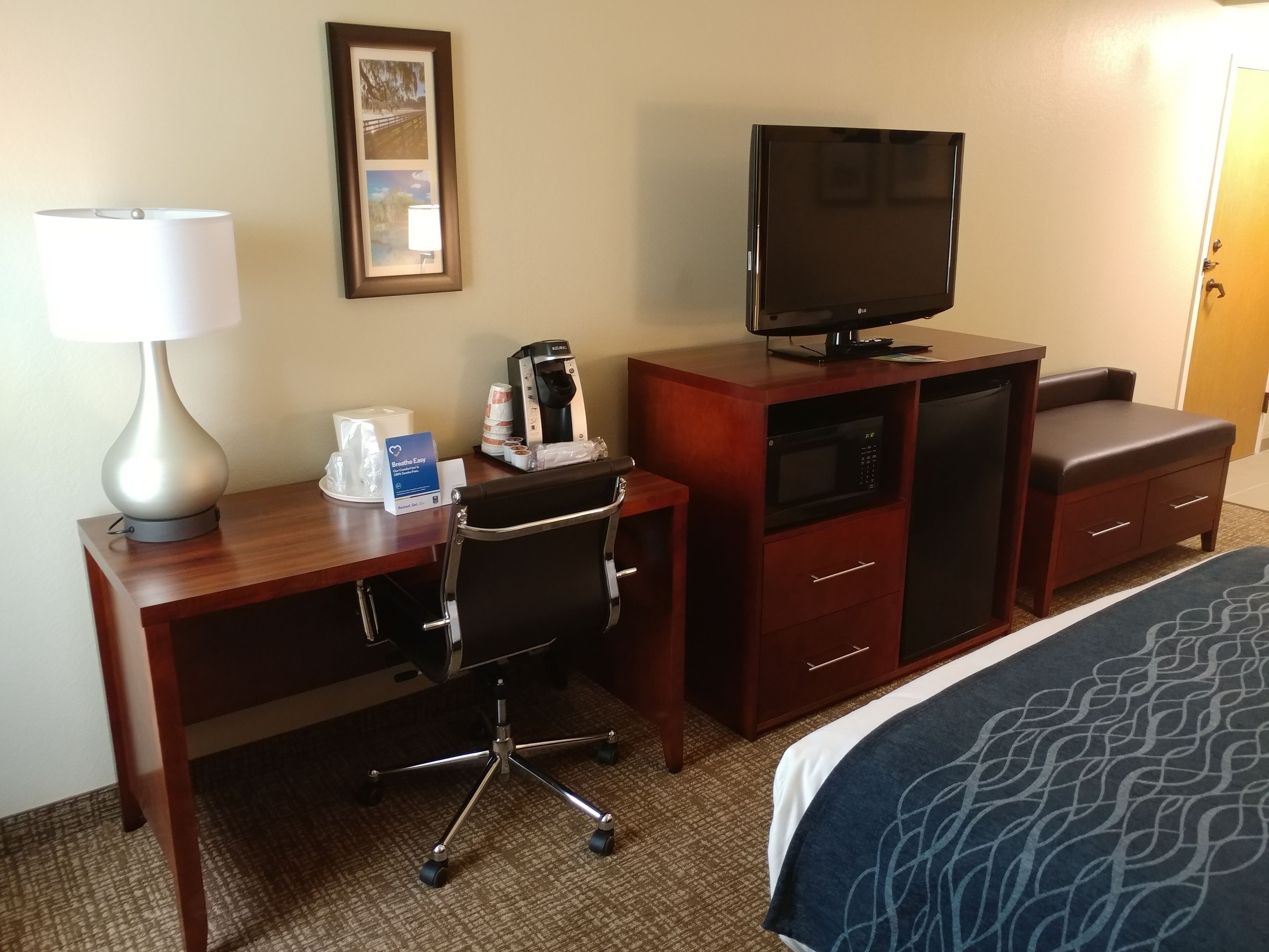 Une chambre d’hôtel avec un bureau en bois, une chaise, une télévision, une machine à café, un four à micro-ondes, un petit placard et un lit recouvert d’une couverture à motifs.