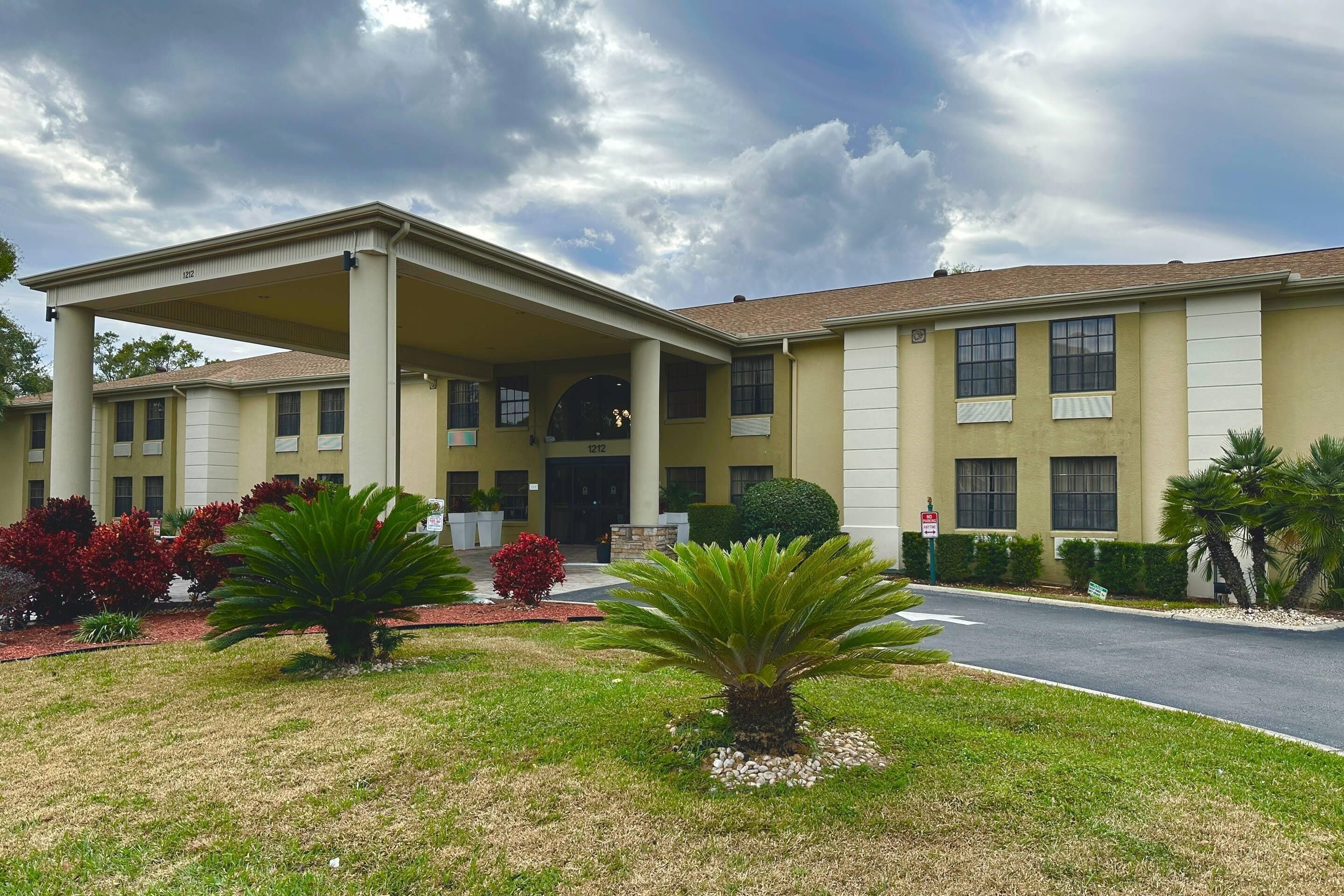 Um hotel de dois andares com entrada coberta, um jardim frontal ajardinado com palmeiras e arbustos, e céu claro.