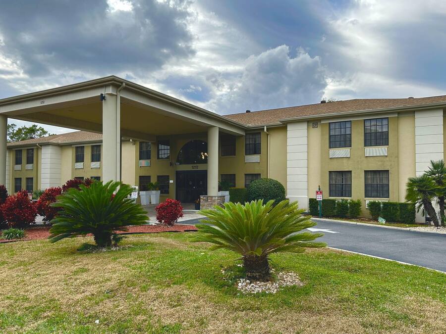 Um hotel de dois andares com entrada coberta, um jardim frontal ajardinado com palmeiras e arbustos, e céu claro.