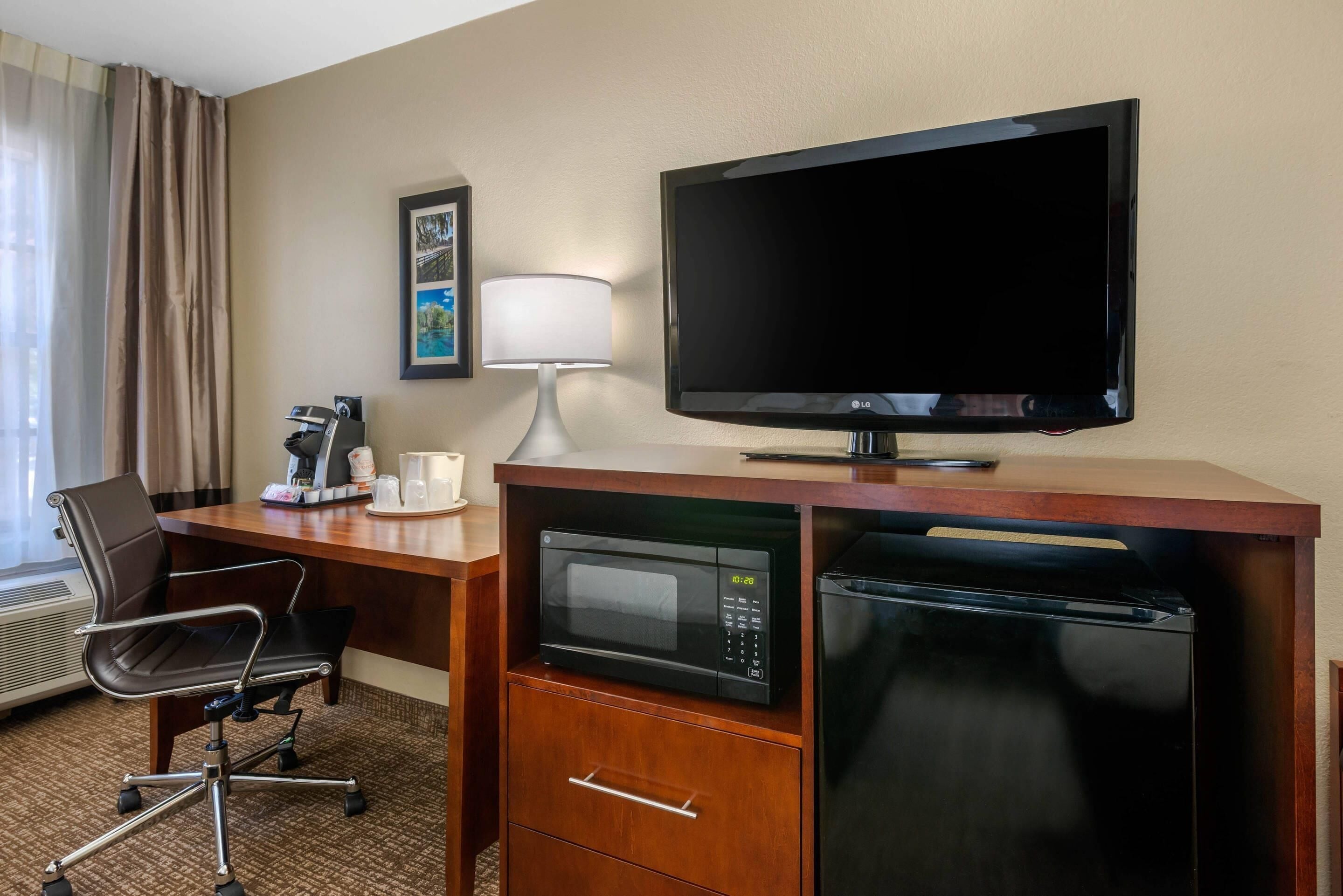 A hotel room with a desk, a television, a microwave, a refrigerator, a chair, a coffee maker, and a framed picture on the wall.