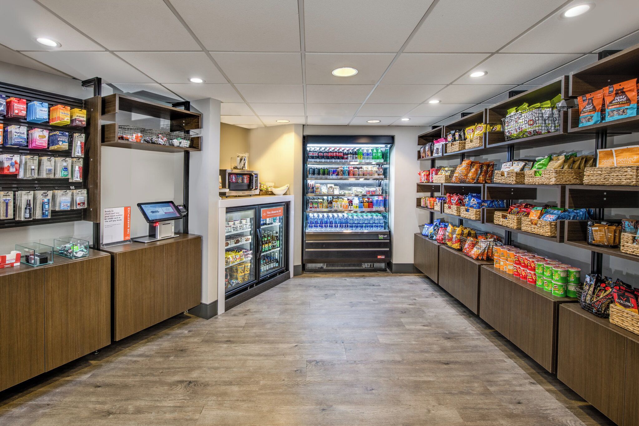 A grocery store with a self-checkout station, refrigerated beverage display, and shelves stocked with snacks and packaged goods.