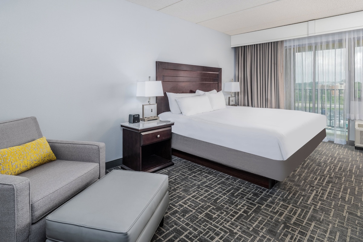 A hotel room with a large bed, a grey sofa, a small table with a lamp, and a window with curtains.
