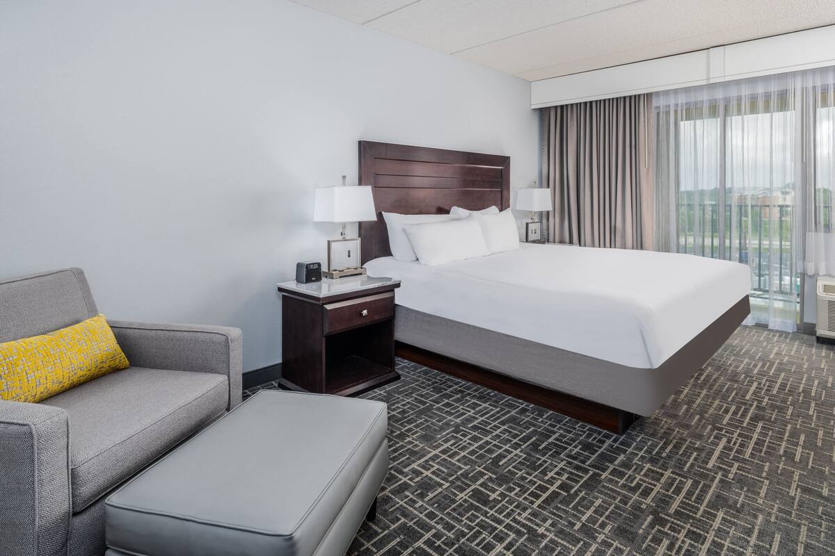Une chambre d’hôtel avec un grand lit, un canapé gris, une petite table avec une lampe et une fenêtre avec des rideaux.
