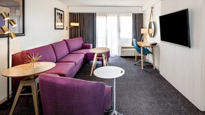 Una habitación de hotel moderna con un sofá morado, dos mesas redondas, un televisor y un ventanal con cortinas.