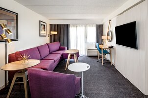 Una moderna camera d'albergo con un divano viola, due tavolini, una TV e un'ampia finestra con tende.