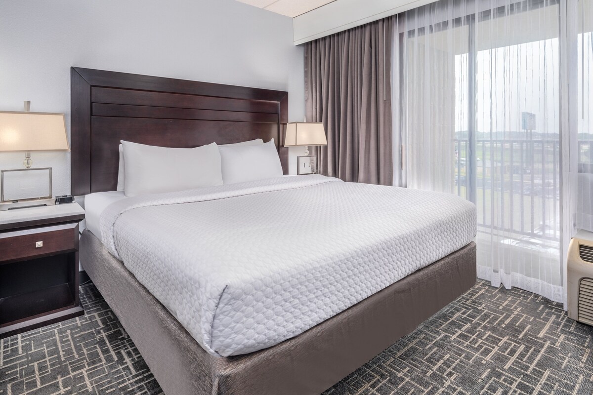 A hotel room with a large bed, two bedside tables with lamps, a window with sheer curtains, and a view of the outside.