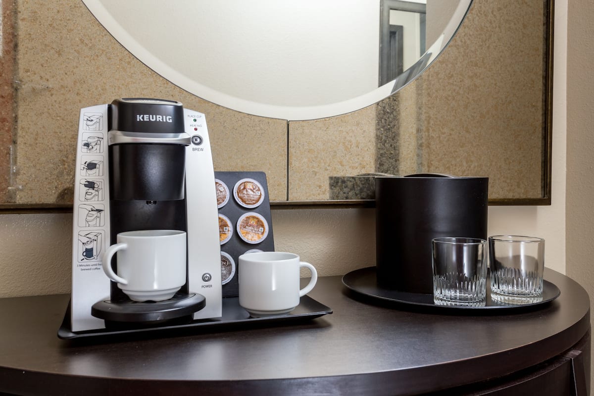 Une machine à café Keurig avec un compartiment pour les dosettes de café, deux tasses blanches et deux verres transparents, le tout posé sur un plateau.