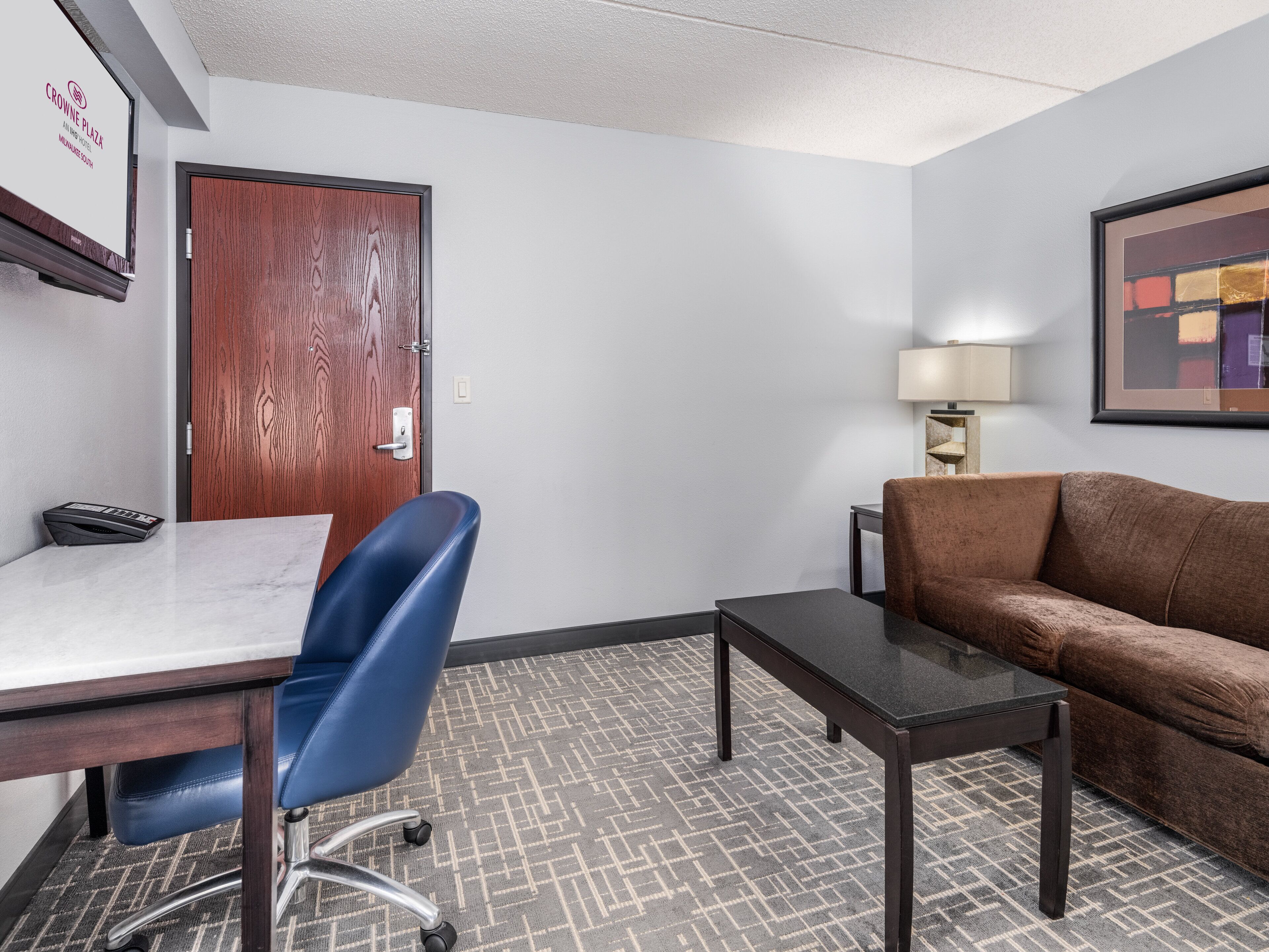 A hotel room with a wooden door, a desk with a chair, a brown sofa, a small table, and a lamp.