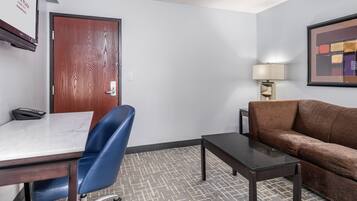 A hotel room with a wooden door, a desk with a chair, a brown sofa, a small table, and a lamp.