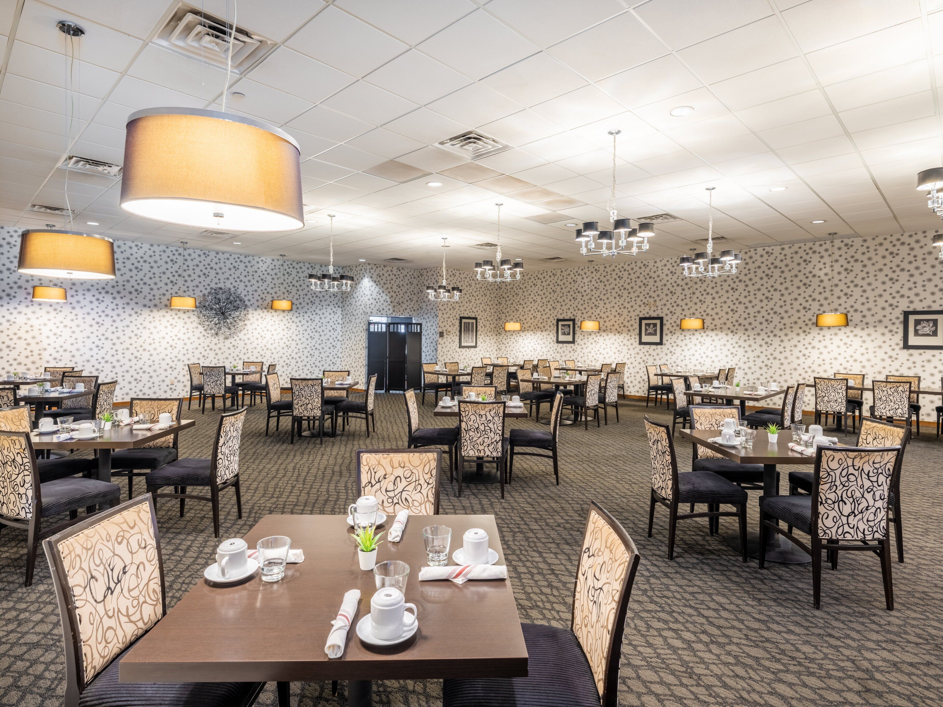 A spacious hotel dining area with multiple tables and chairs, patterned wallpaper, and pendant lighting.