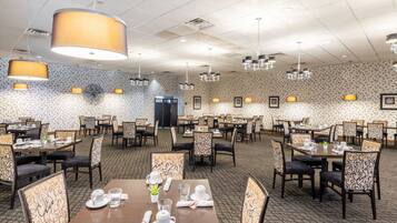 A spacious hotel dining area with multiple tables and chairs, patterned wallpaper, and pendant lighting.