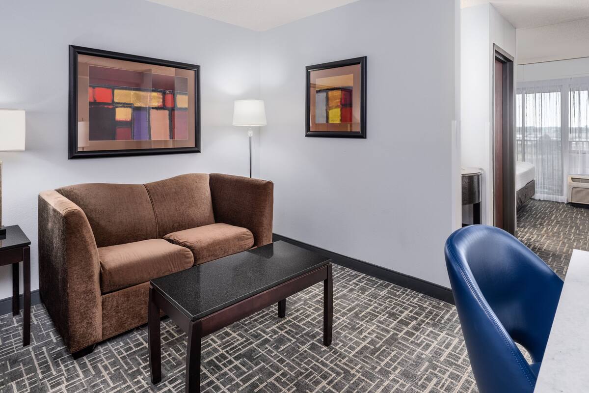 Une chambre d’hôtel avec un canapé marron, une petite table, une chaise bleue et deux tableaux abstraits encadrés accrochés au mur.