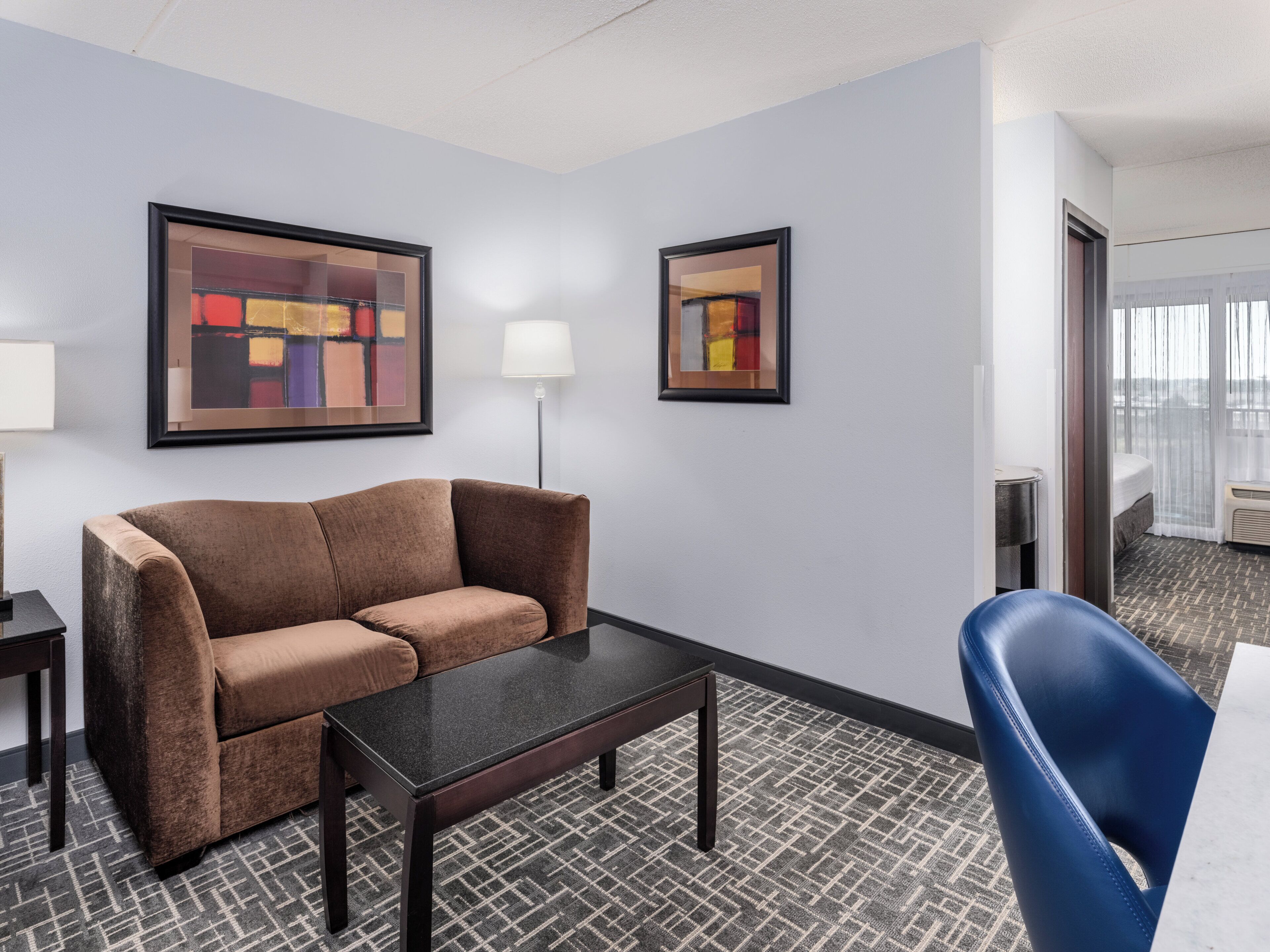 A hotel room with a brown sofa, a small table, a blue chair, and two framed abstract paintings on the walls.