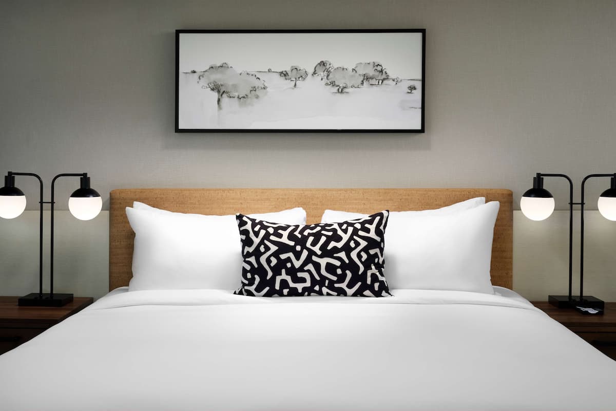 A neatly made bed with white and patterned pillows, a framed artwork above it, and two bedside lamps on either side.