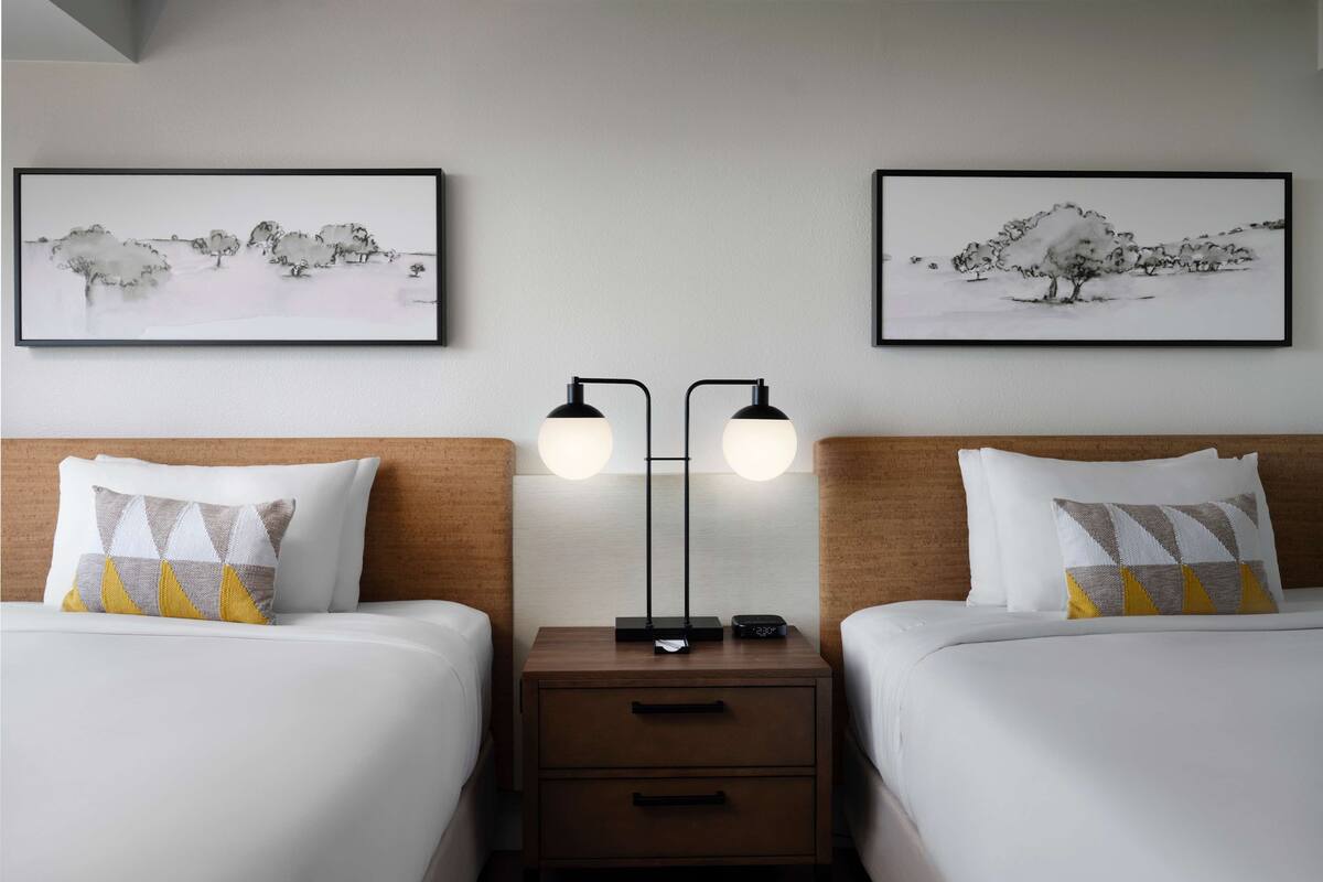 A hotel room with two beds, each with a wooden headboard, white bedding, and patterned pillows. There are two bedside tables with lamps and a small drawer. Two framed artworks depicting trees are hung on the wall above the beds.