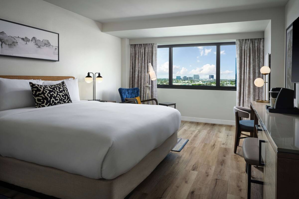 A hotel room with a large bed, a desk with a chair, a view of the city through the window, and a painting on the wall.