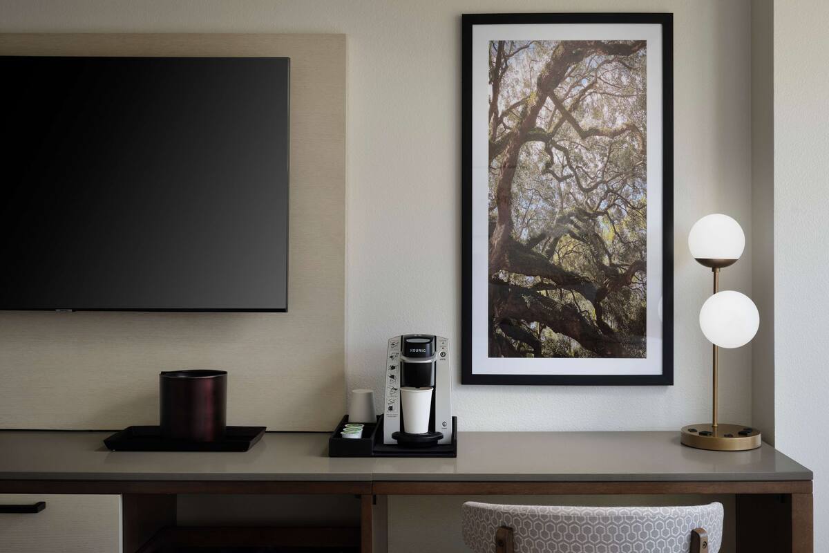 A modern hotel room with a flat-screen TV, a framed tree photograph, a coffee maker, and a two-lamp floor lamp.