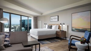 Habitación de hotel moderna con una cama grande, un sofá, una silla y un escritorio. Se ve una ventana con vista a la ciudad.