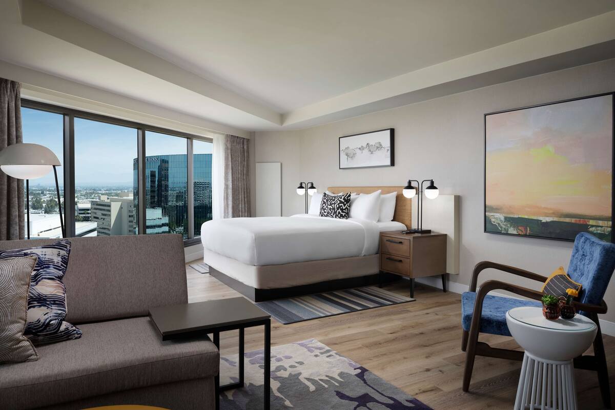 A modern hotel room with a large bed, a sofa, a chair, and a desk. There is a window with a city view.