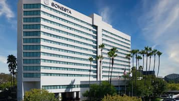 A modern hotel building with the name 'SONESTA' on the facade, featuring a row of windows and a parking area with several cars and palm trees.