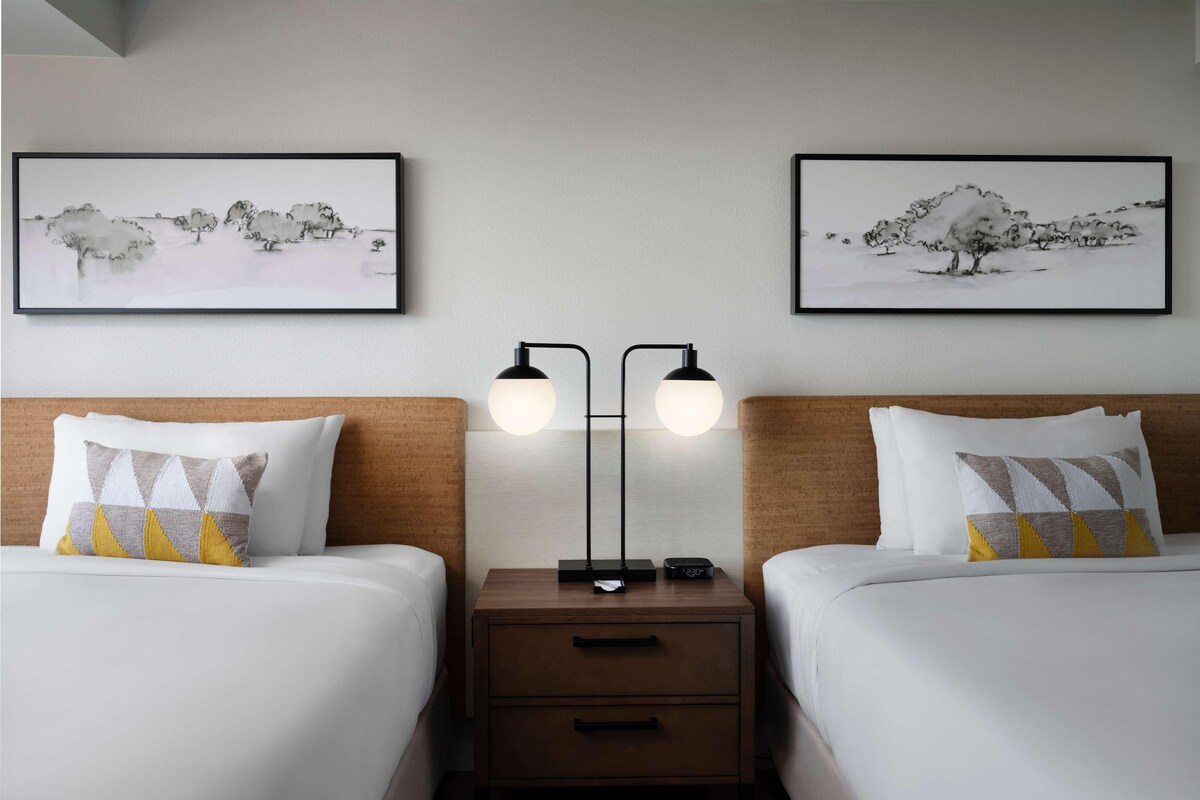 Une chambre d’hôtel avec deux lits, une table de chevet, une lampe et deux tableaux encadrés au mur.