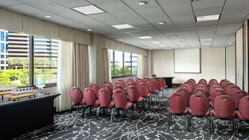 A conference room with rows of red patterned chairs, a projector screen, a presentation table, and a large window with curtains.