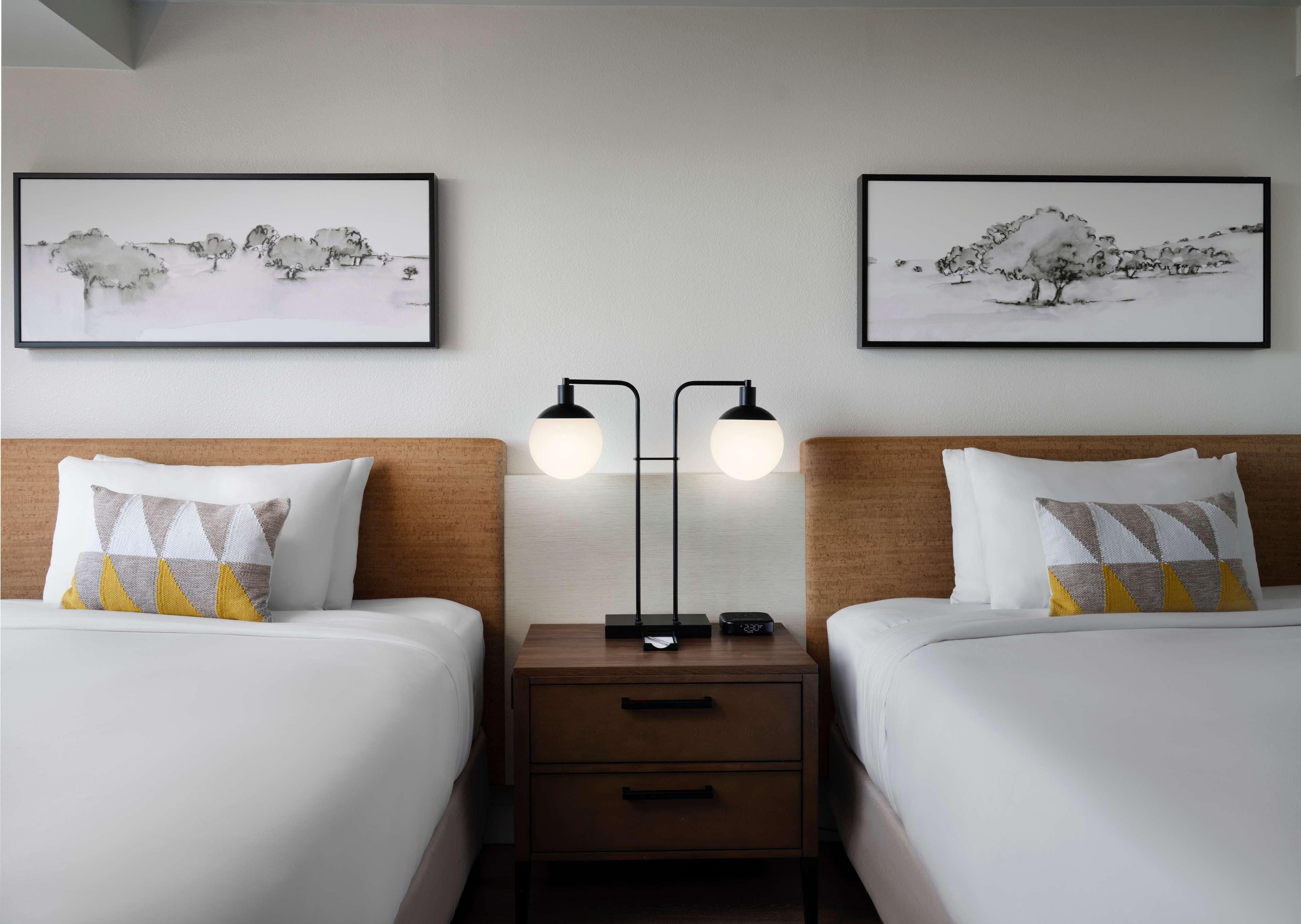Une chambre d’hôtel avec deux lits, une table de chevet, une lampe et deux tableaux encadrés au mur.