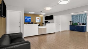 A hotel reception area with a reception desk, a black leather sofa, a kitchen area, and a wall-mounted TV.