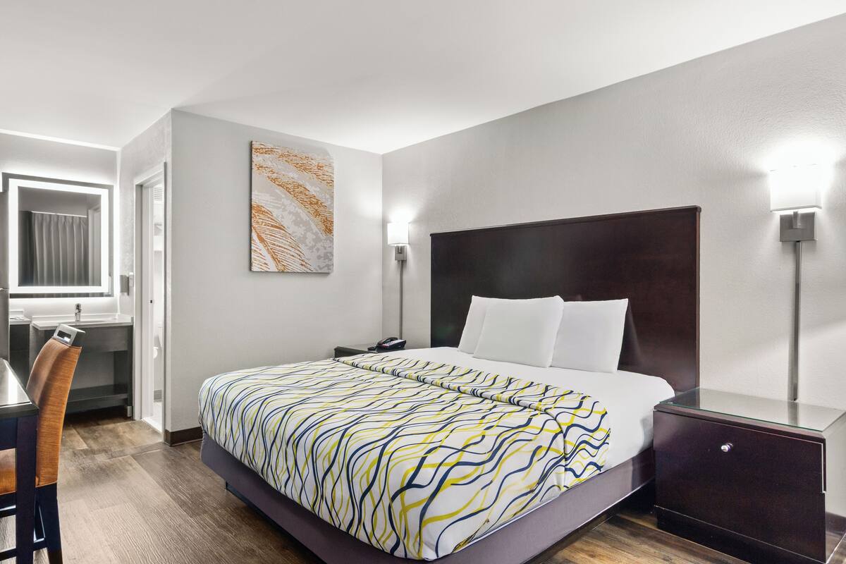 A hotel room with a bed, bedside table, lamp, and a painting on the wall.