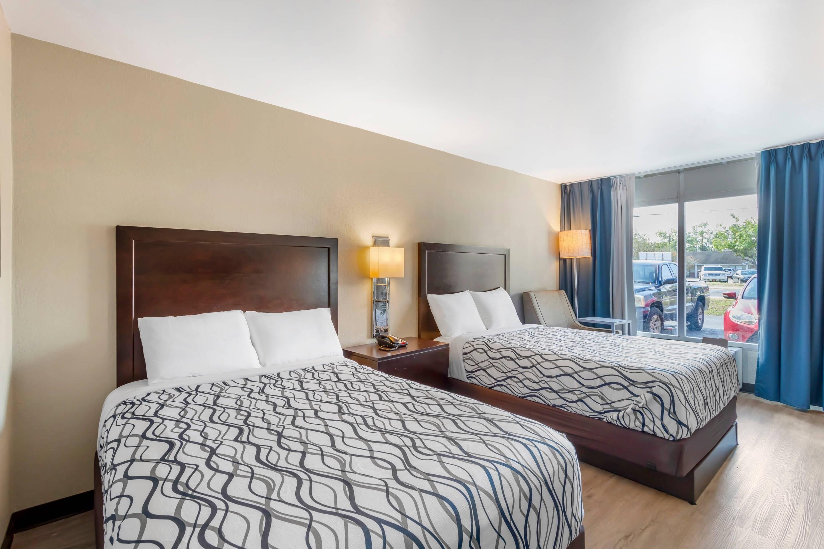 A hotel room with two beds, a nightstand with a lamp, a window with curtains, and a view of parked cars outside.