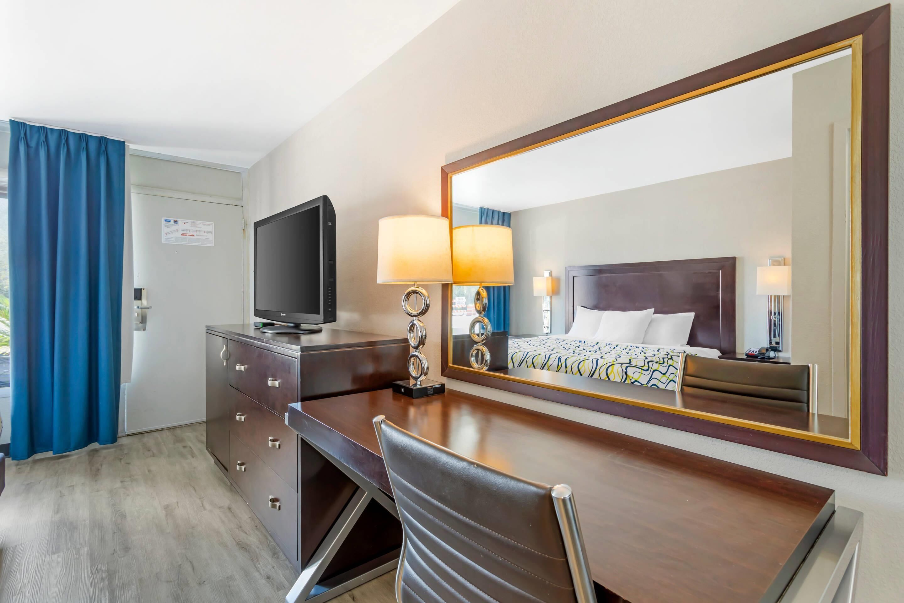 Een hotelkamer met een bed, een televisie, een bureau met lades, een stoel en een grote spiegel.