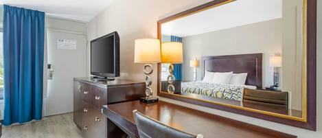 Habitación de hotel con cama, televisión, escritorio con cajones, silla y espejo grande.