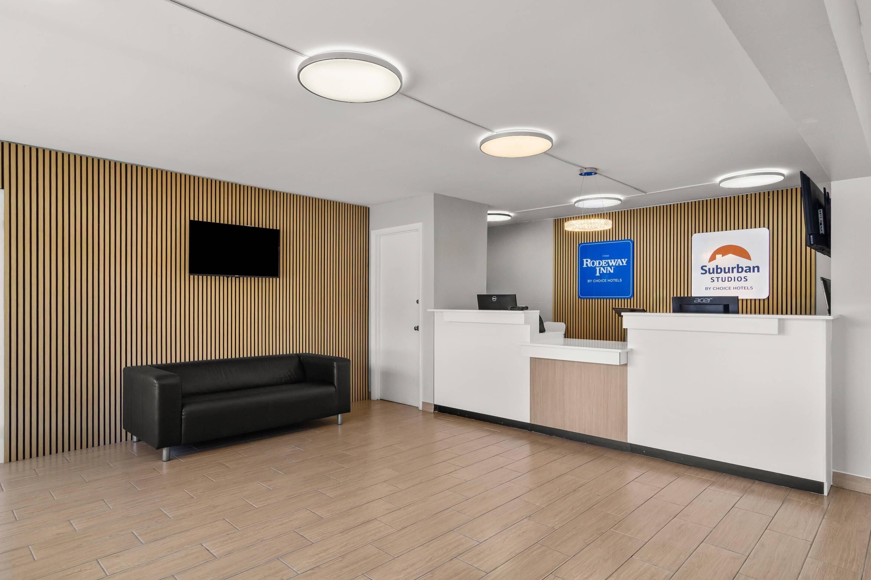 A reception area with a black sofa, wooden paneling, a reception desk, and signs for 'RODEWAY INN' and 'Suburban Studios.'