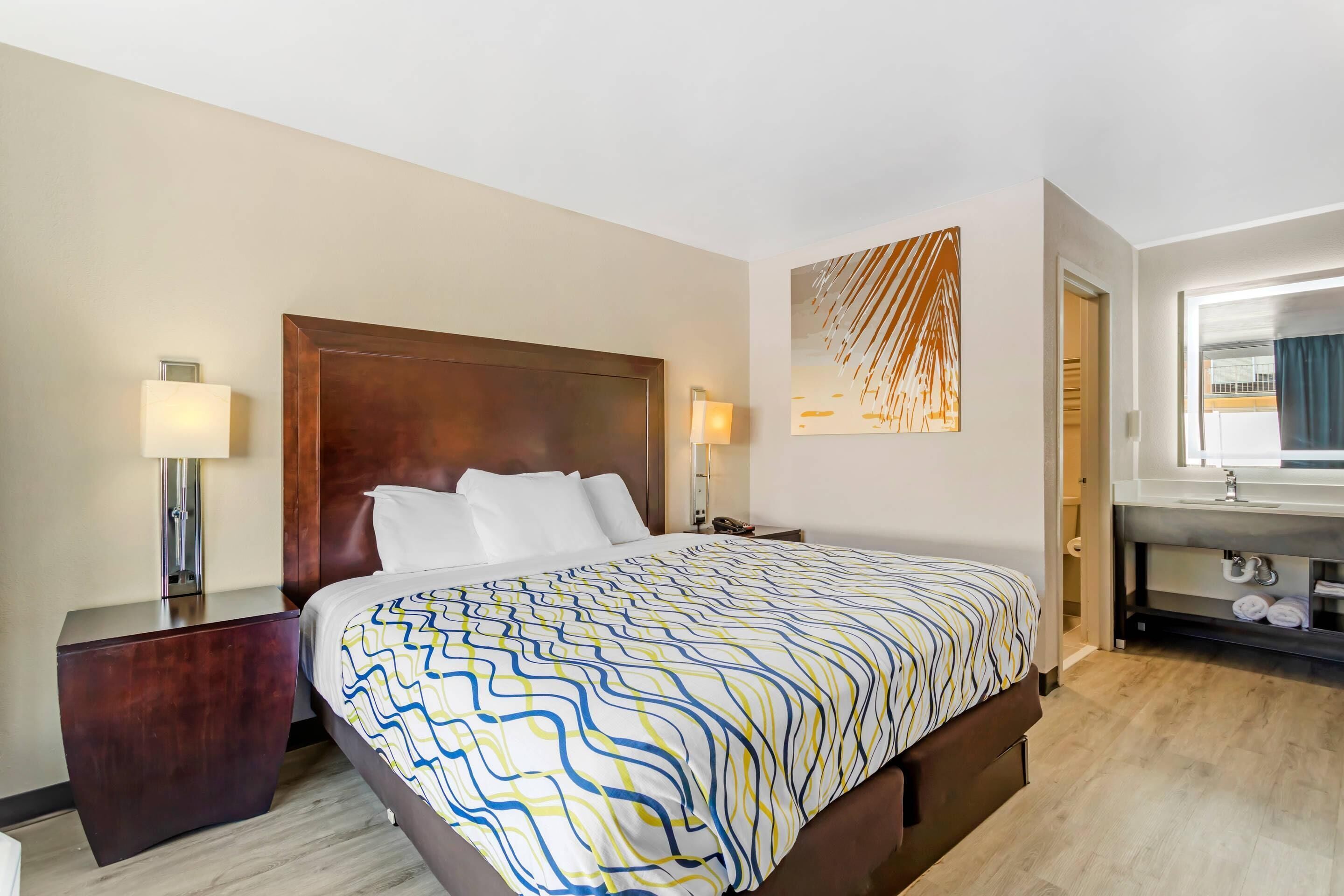 Ein Hotelzimmer mit einem großen Bett, Nachttischlampen, einem Holzkopfteil, einem Wandbild und einem Badezimmer mit Waschbecken und Handtuchhalter.