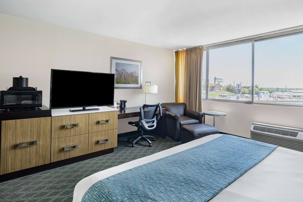 Ein Hotelzimmer mit Bett, Schreibtisch, Flachbildfernseher, Mikrowelle, Sessel, Lampe und einem großen Fenster mit Blick auf die Stadt.