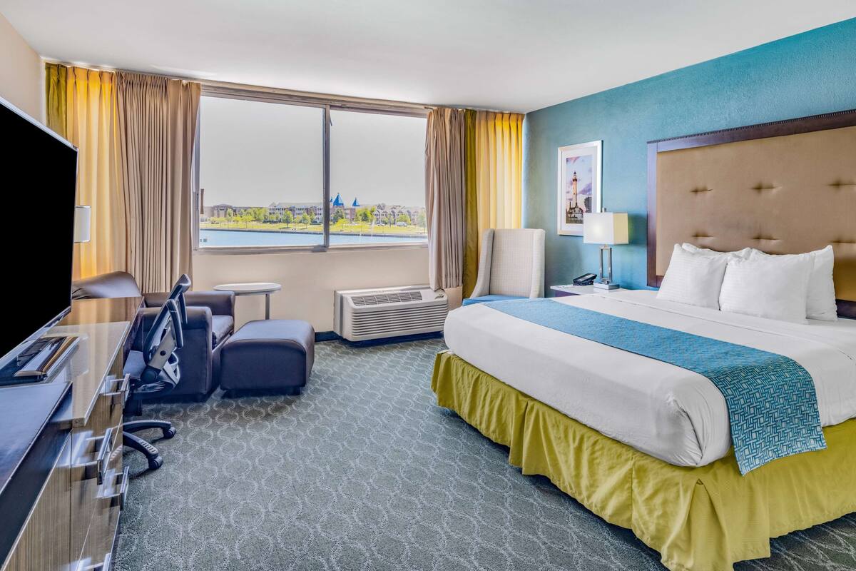 Ein Hotelzimmer mit einem großen Bett, einem Schreibtisch, einem Stuhl, einem Fernseher und einem Fenster mit Blick auf ein Gewässer und Gebäude.