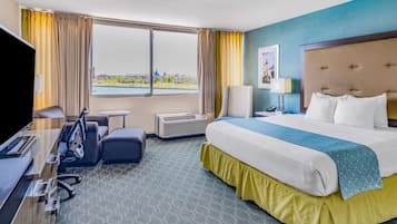 Habitación de hotel con una cama grande, un escritorio, una silla, un televisor y una ventana con vistas a un cuerpo de agua y edificios.