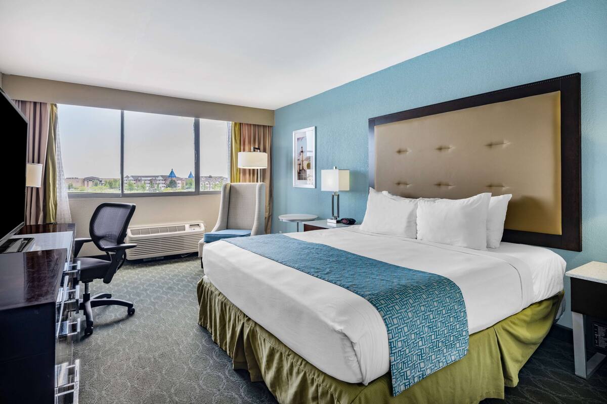 Ein Hotelzimmer mit einem großen Bett, einem Schreibtisch, einem Sessel und Blick auf Gebäude durch das Fenster.