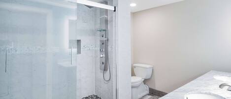 Un baño moderno con ducha envuelta en vidrio, un inodoro blanco y un encimera de mármol con dos lavamanos.