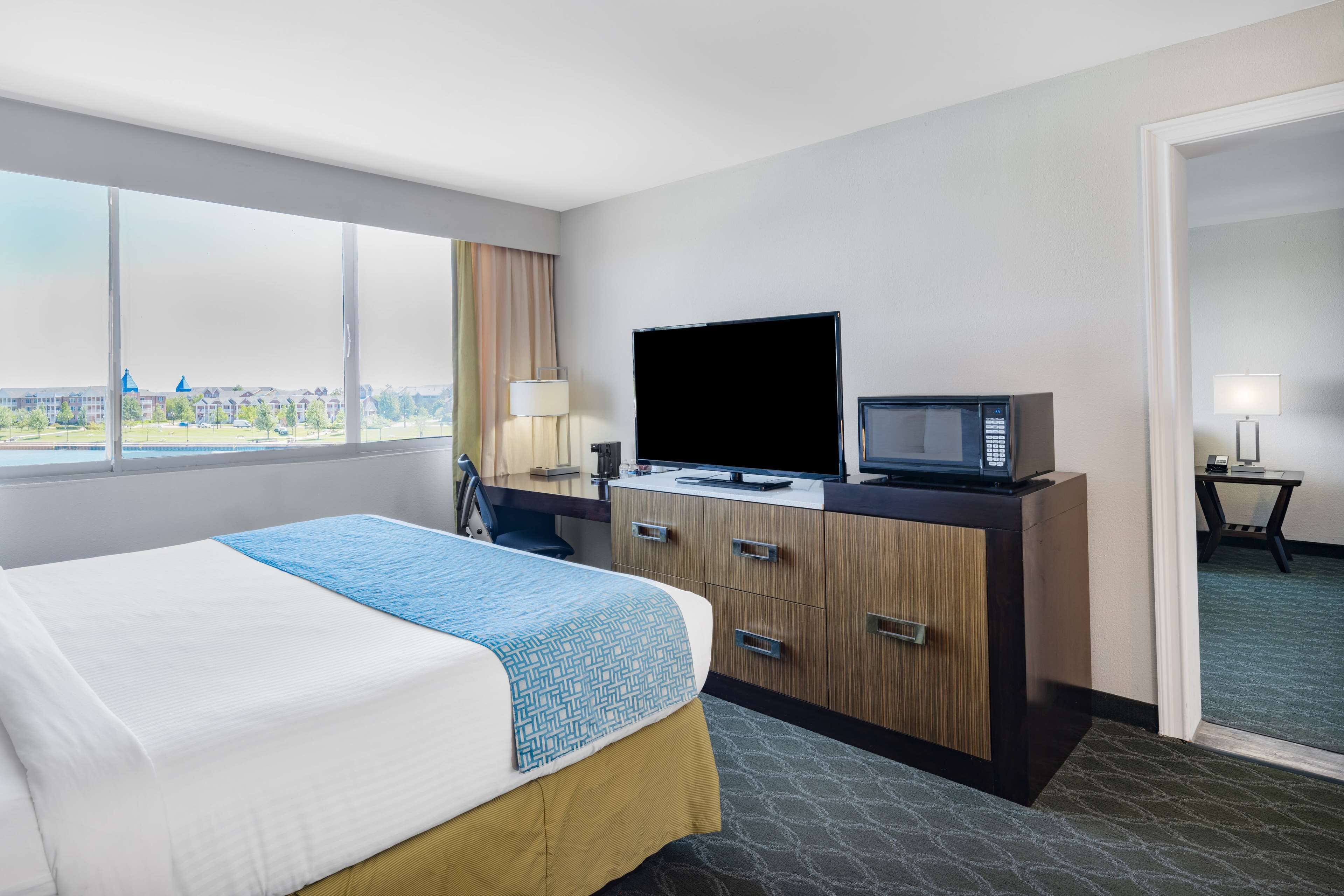A hotel room with a bed, a desk with a TV and a lamp, and a view of buildings outside.