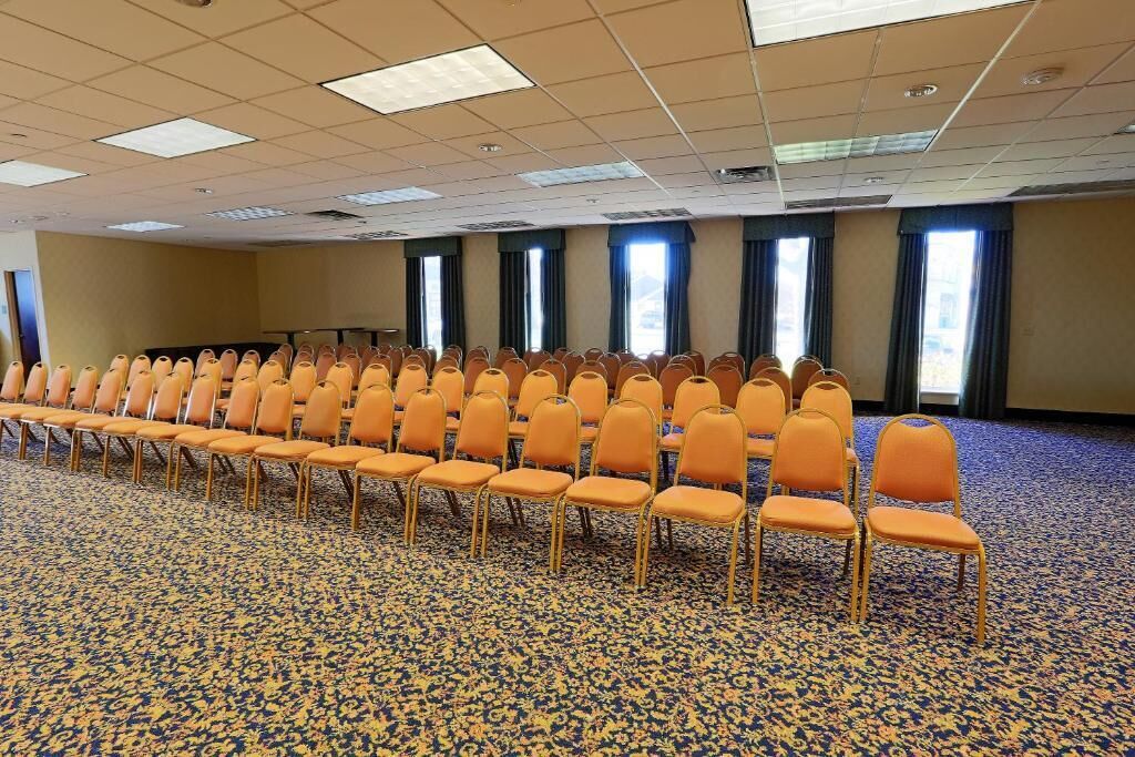 Un salón de conferencias con filas de sillas anaranjadas, suelo alfombrado y ventanas amplias con cortinas.
