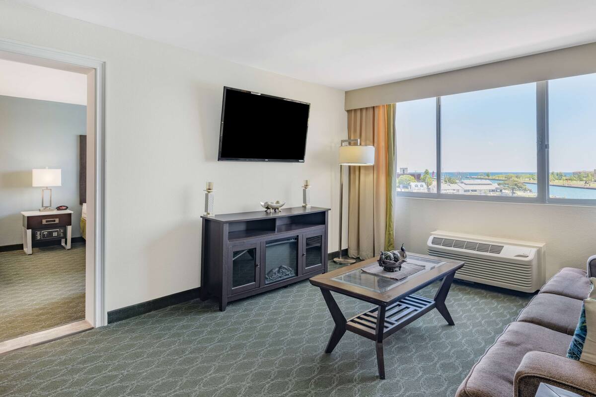 Ein Hotelzimmer mit einem wandmontierten Flachbildfernseher, einer dunklen Holzunterhaltungsecke, einem Glastisch, einem braunen Sofa und einem großen Fenster mit Blick auf das Meer und die Stadt.