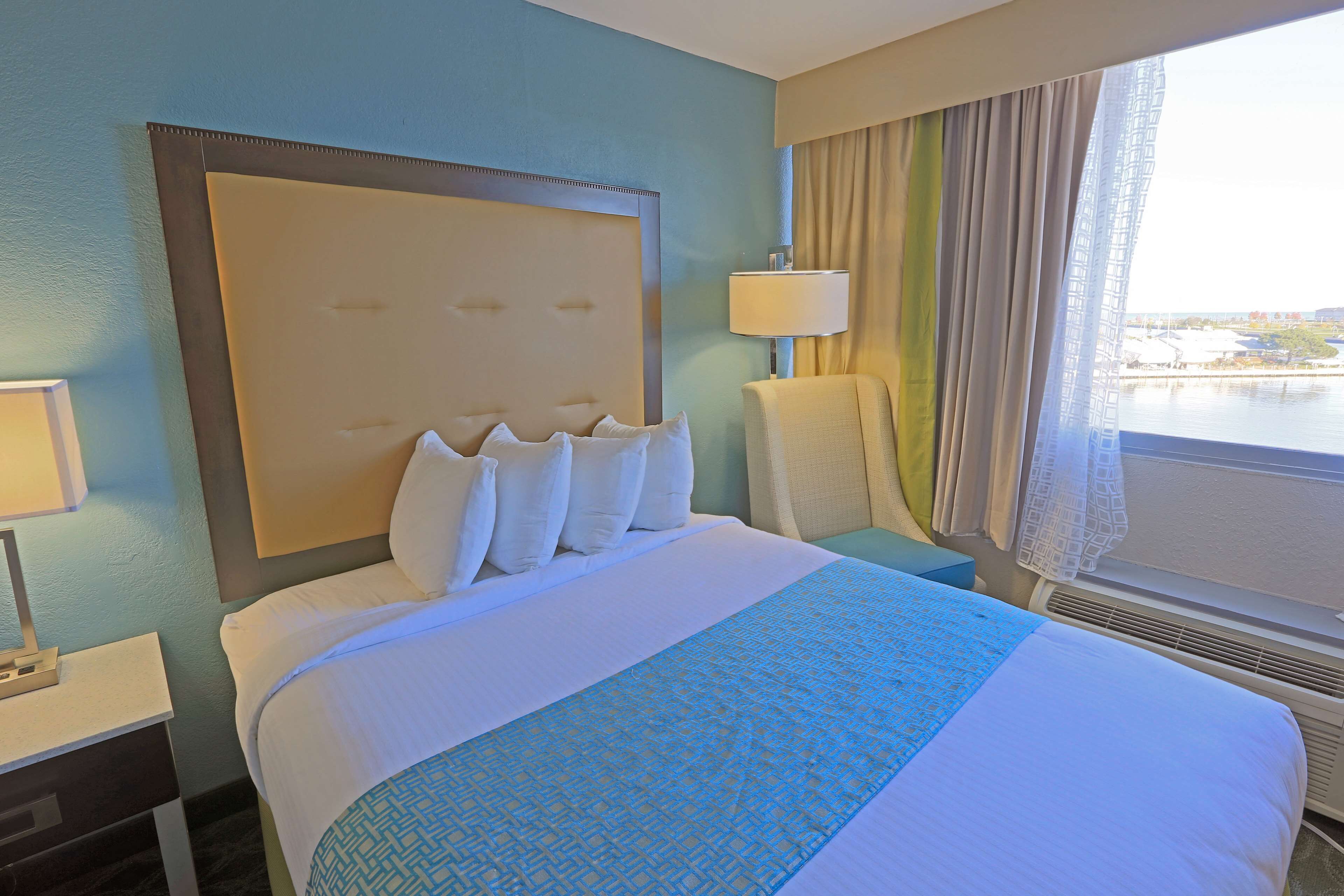 Habitación de hotel con una cama grande, una mesita de noche con lámpara, una silla y vista al agua a través de la ventana.